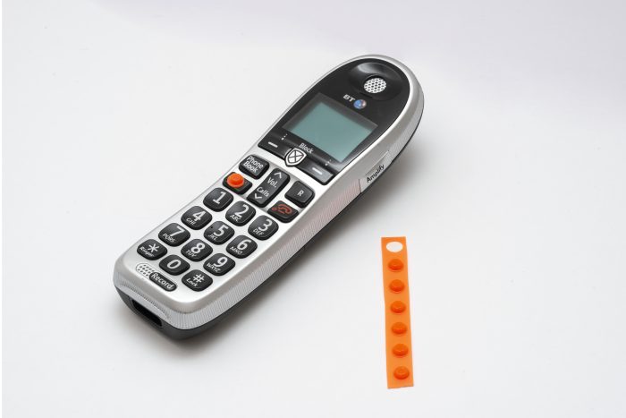 A phone which has an orange tactile sticker known as a bumpon attached to the answer button. Next to it is a strip of the bumpons.