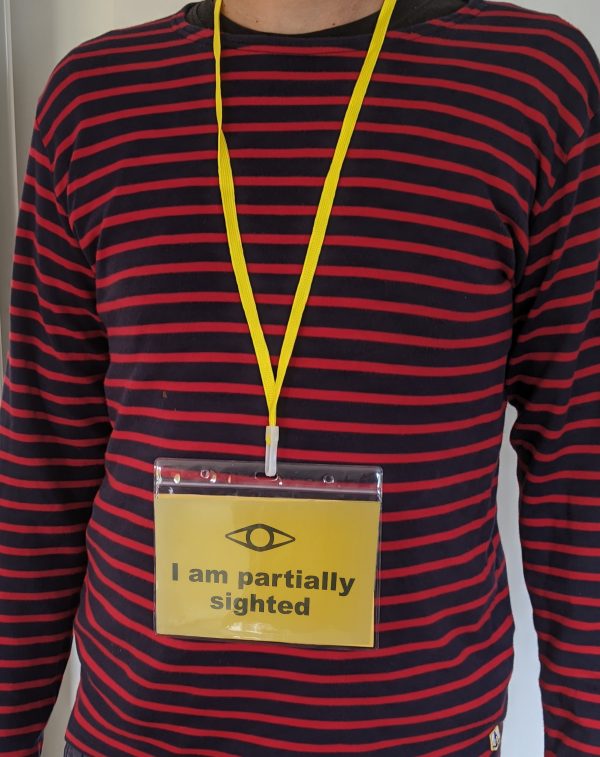 A yellow rectangle badge with the words I am partially sighted sits in a clear plastic wallet hanging from someon's neck by a bright yellow lanyard